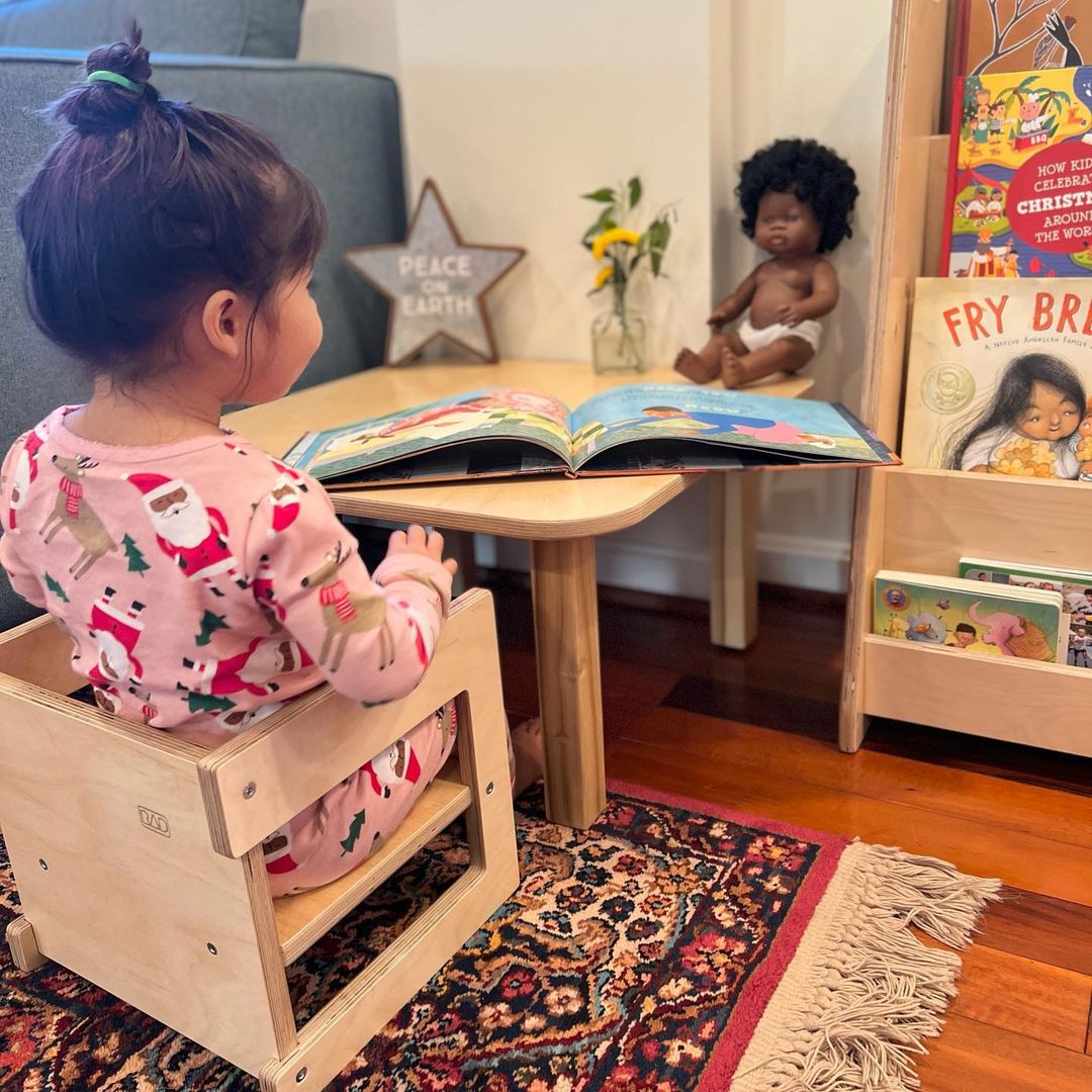 Child sitting on a wooden stool reading a book in a home setting with toys and books around.