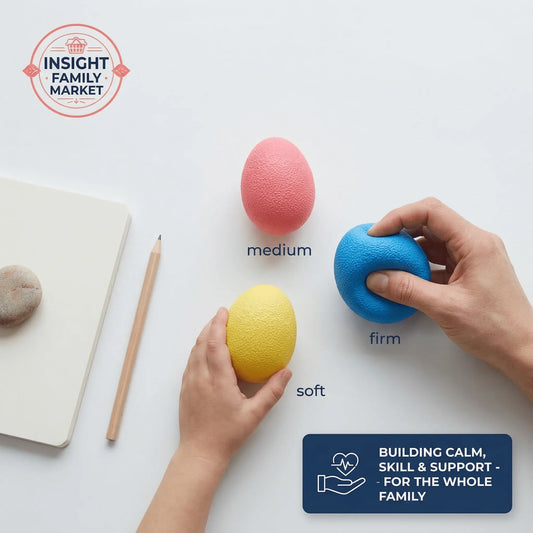 Three textured egg-shaped hand exercise stress balls in blue, pink, and yellow, labeled firm, medium, soft, held by adult and child hands on a white desk.