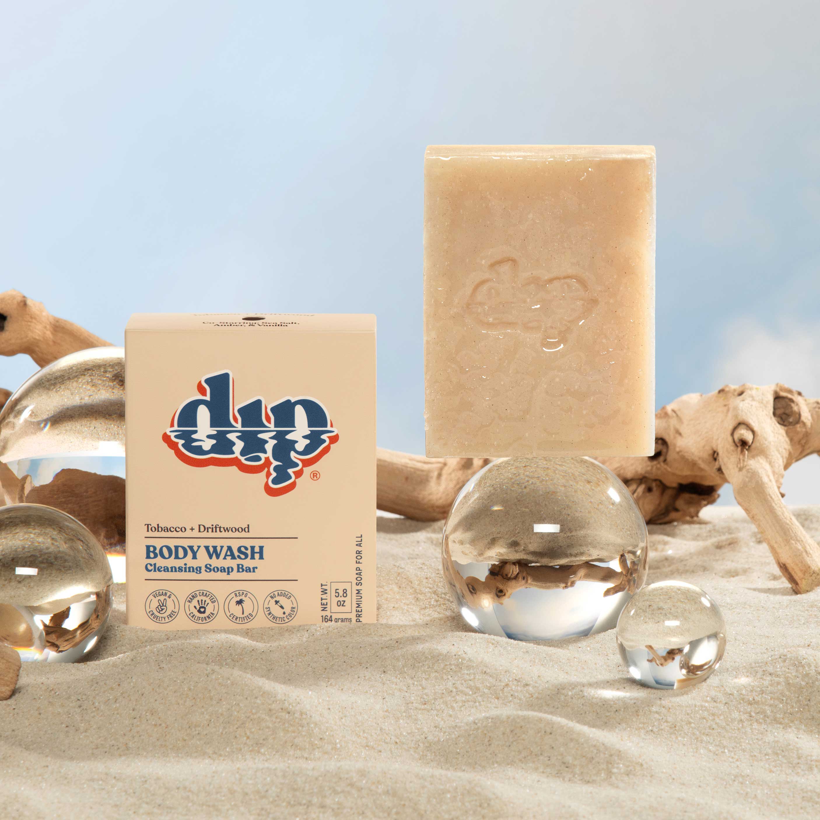 Dip body wash bar and packaging on a sandy surface with glass spheres and driftwood.