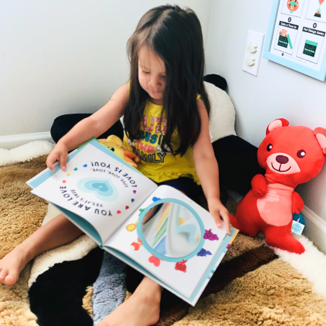 Child reading a book with a red teddy bear toy next to them on a carpeted floor.