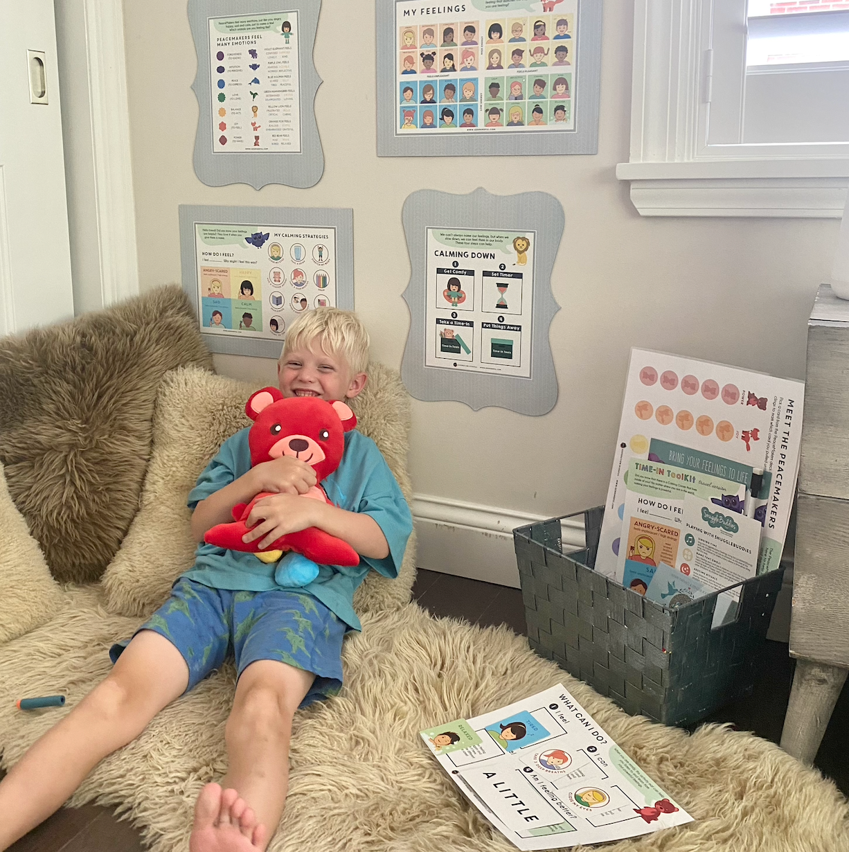 Child sitting on a couch holding a red teddy bear with educational posters on the wall.