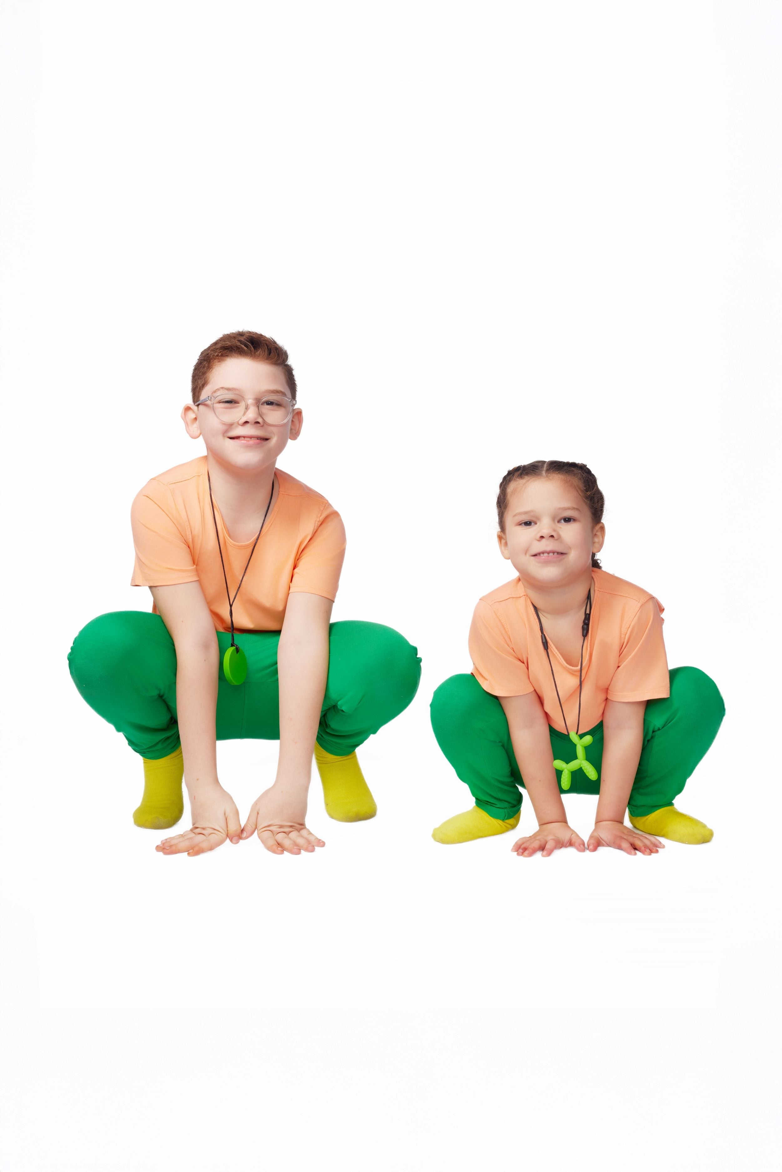 Two children in matching outfits with green pants and orange tops on a white background