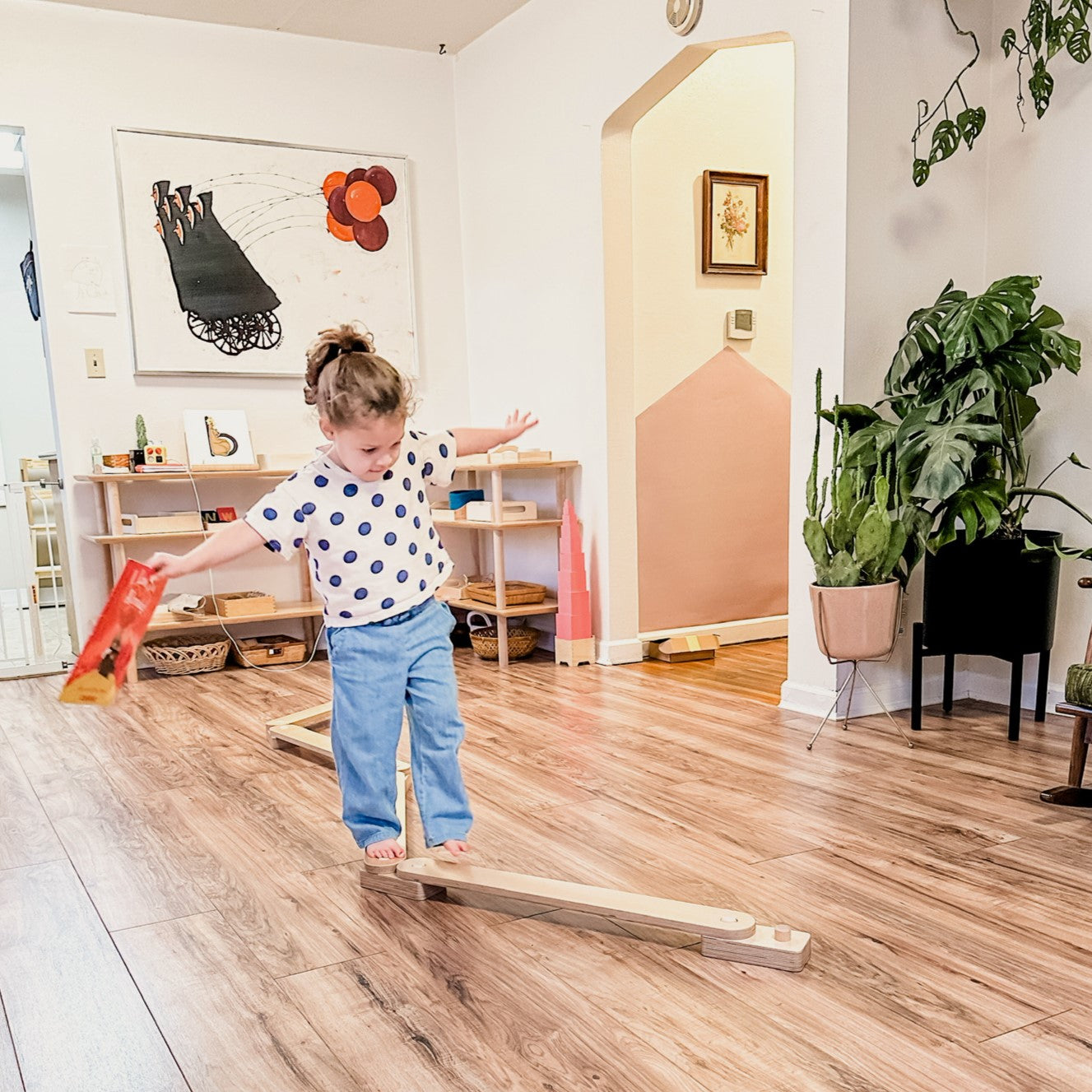 Child balancing on a wooden seesaw in a home setting with plants and furniture.