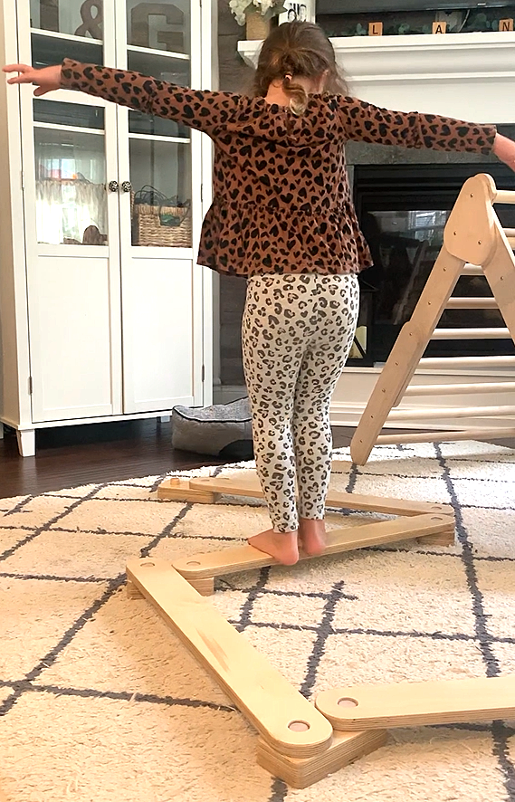 Person wearing a leopard print outfit standing on a wooden balance beam in a home setting.