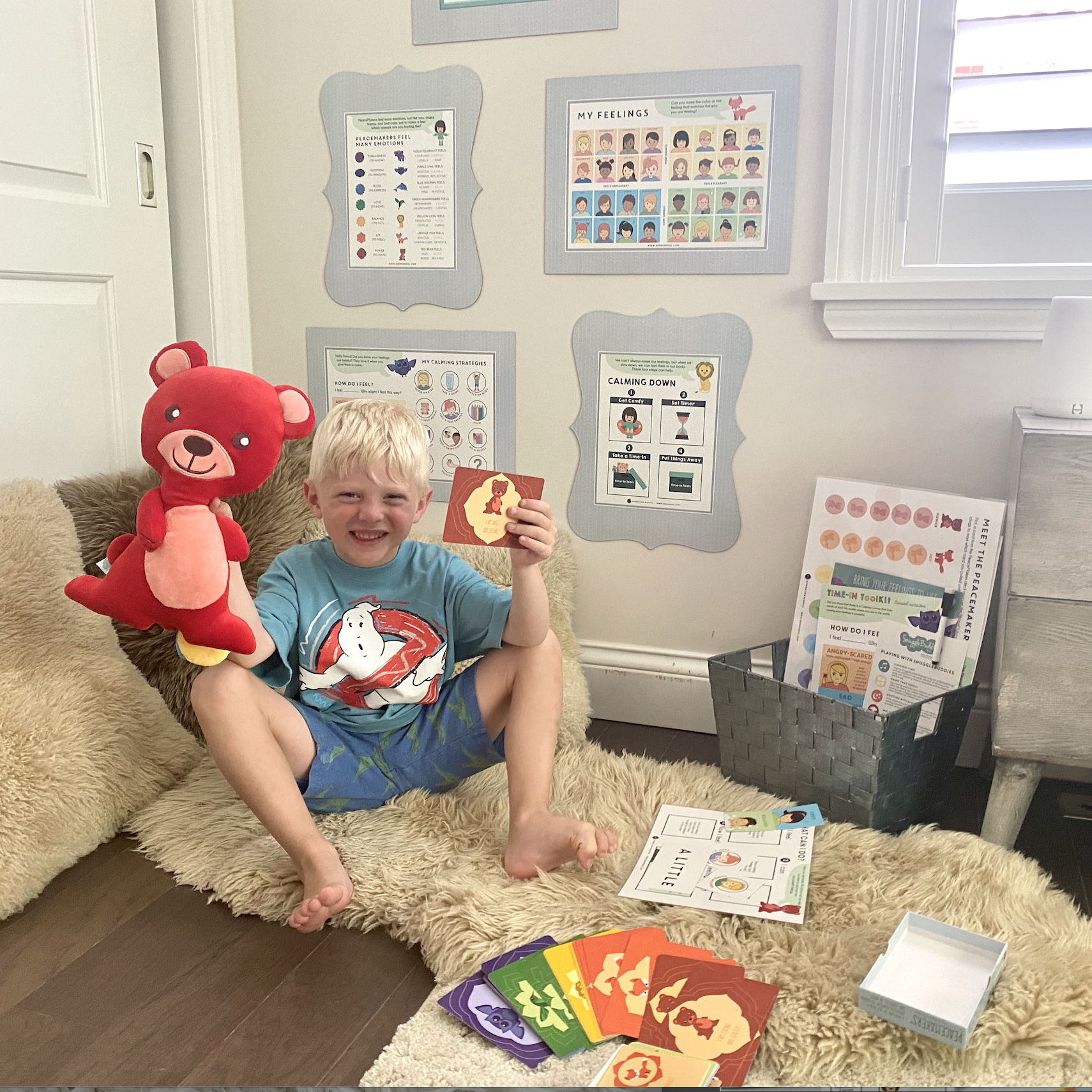 Child sitting on a couch with a teddy bear, holding a card, in a room with educational posters on the wall.