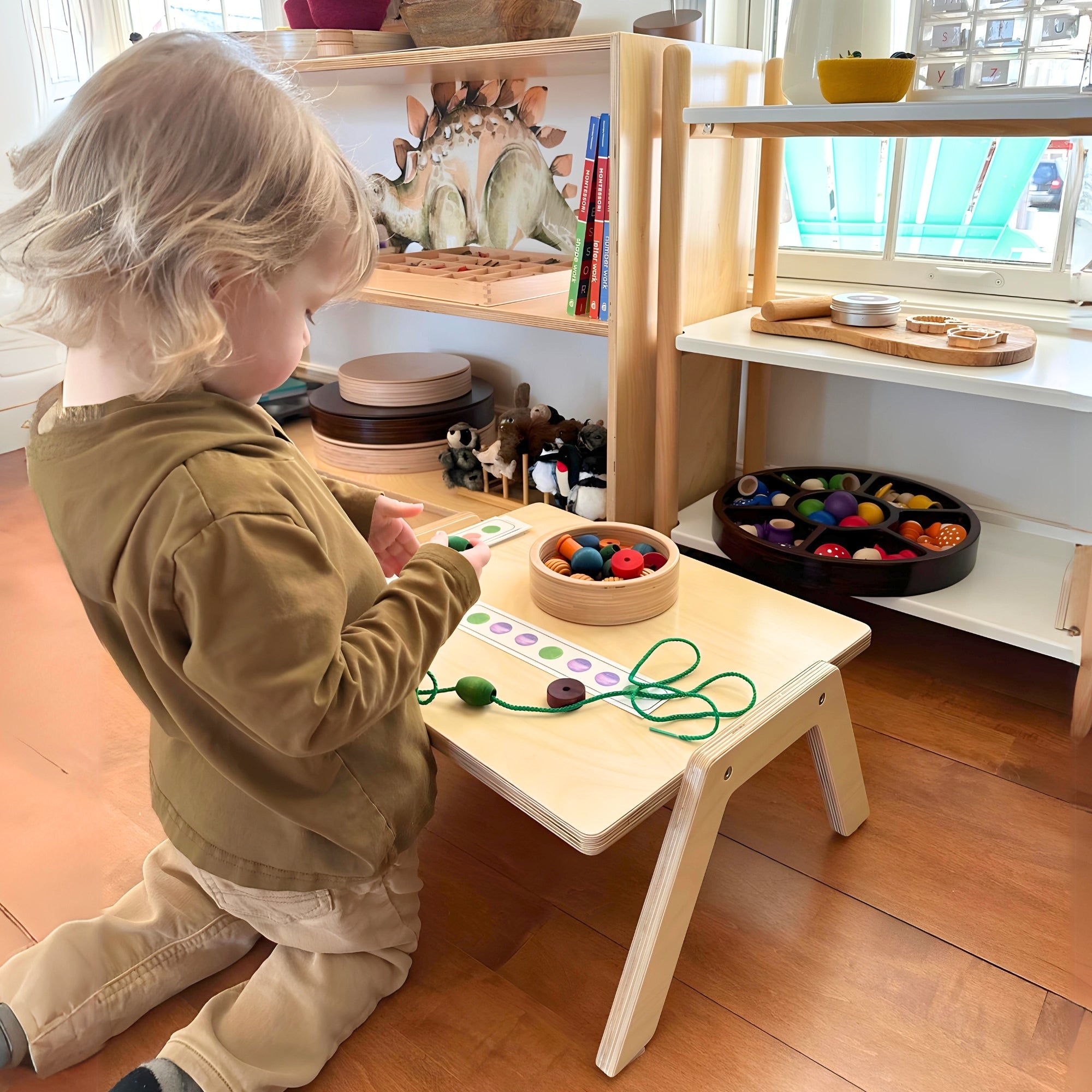 Child playing with educational toys at a wooden table in a room with shelves and windows.