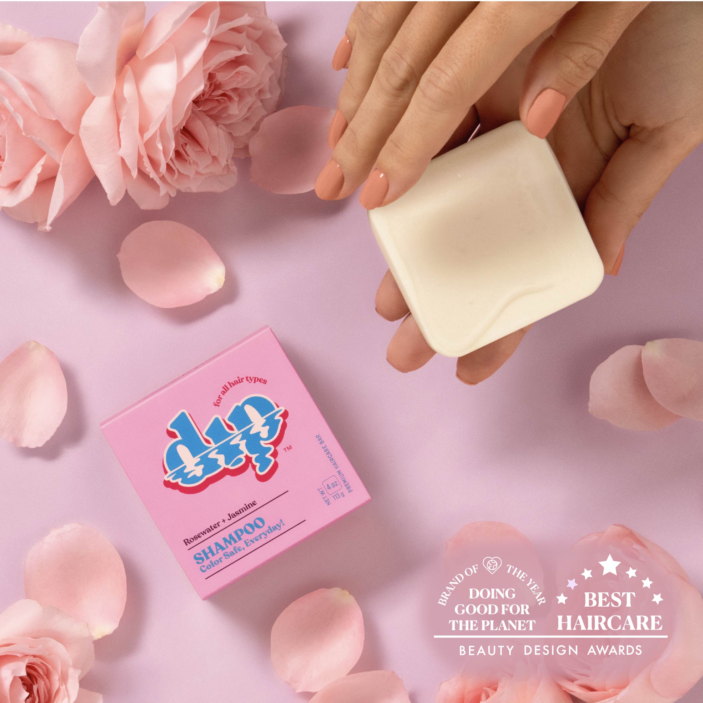 Hand holding a bar of soap with a pink shampoo box and roses on a pink background