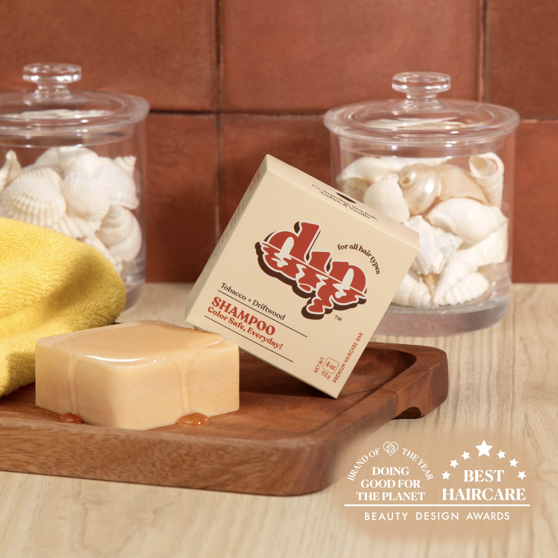 Bar of soap and shampoo bar with a label on a wooden tray, surrounded by jars and a yellow sponge.