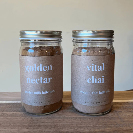 Two glass jars of latte mixes on a wooden surface, labeled golden nectar golden milk latte mix and vital chai cacao chai latte mix.
