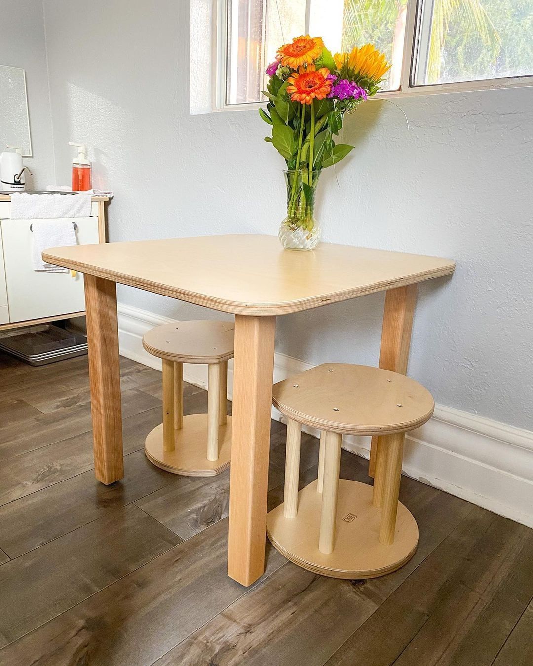 Wooden dining table with three stools in a room with a window and kitchen area.