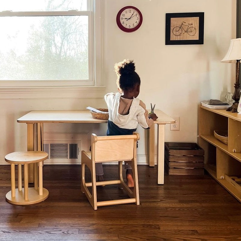 Child sitting at a small wooden table in a room with a window and furniture.