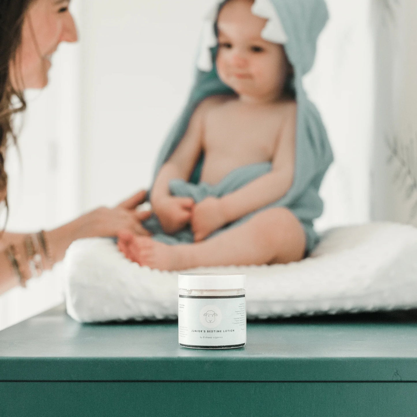 Bedtime lotion jar on table, baby in hooded towel sitting on changing pad, soft lighting