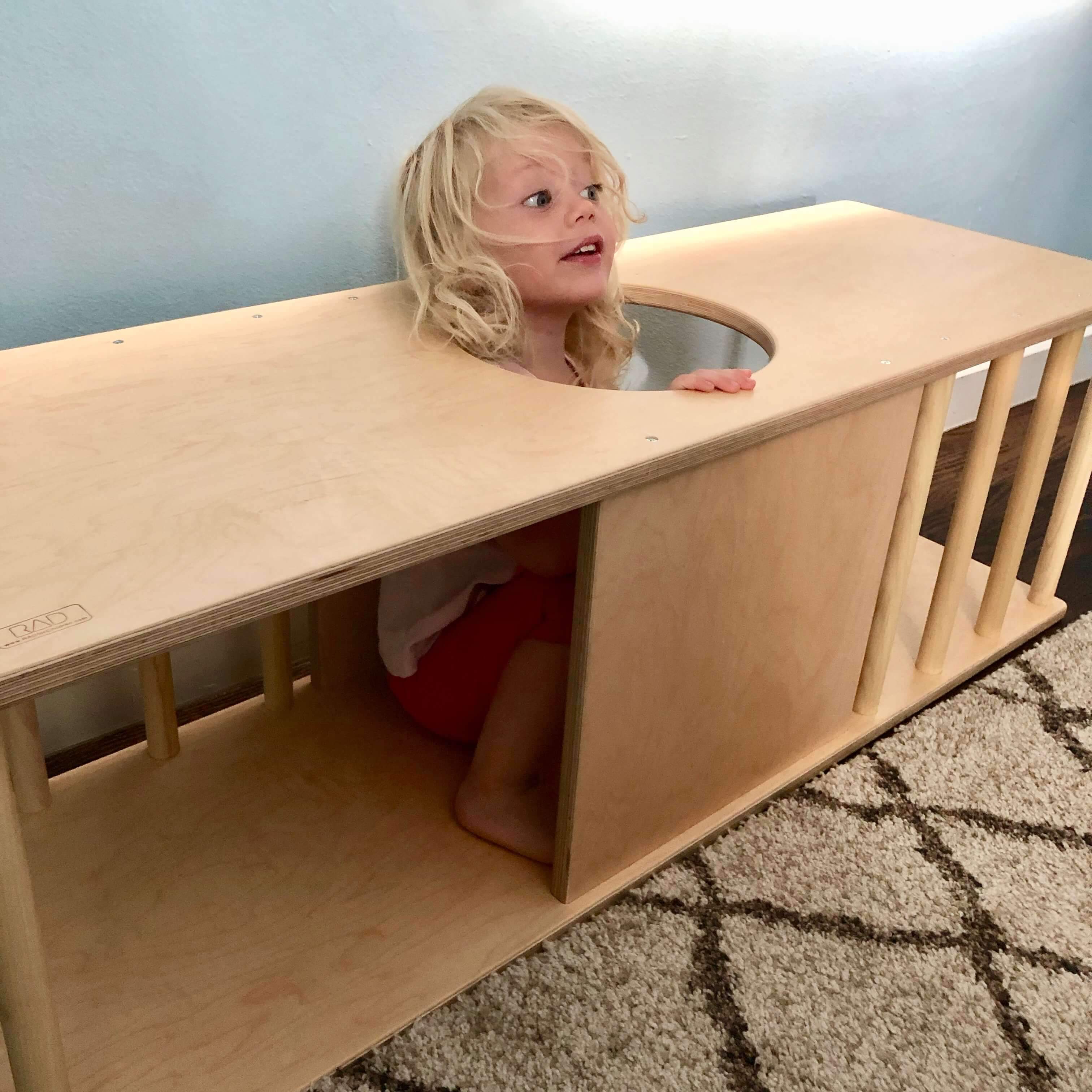 Child playing inside a wooden toy structure with a light blue wall in the background