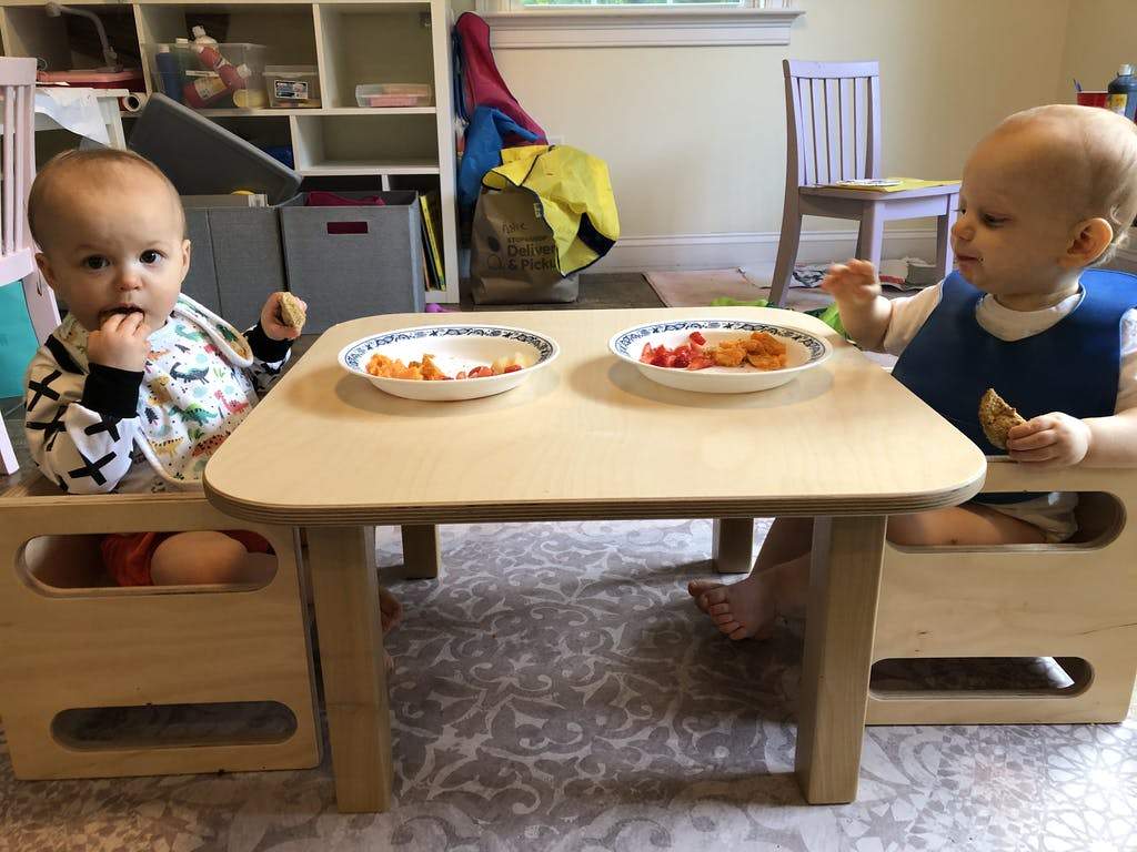 Two children sitting at a small wooden table with plates of food in a home setting.