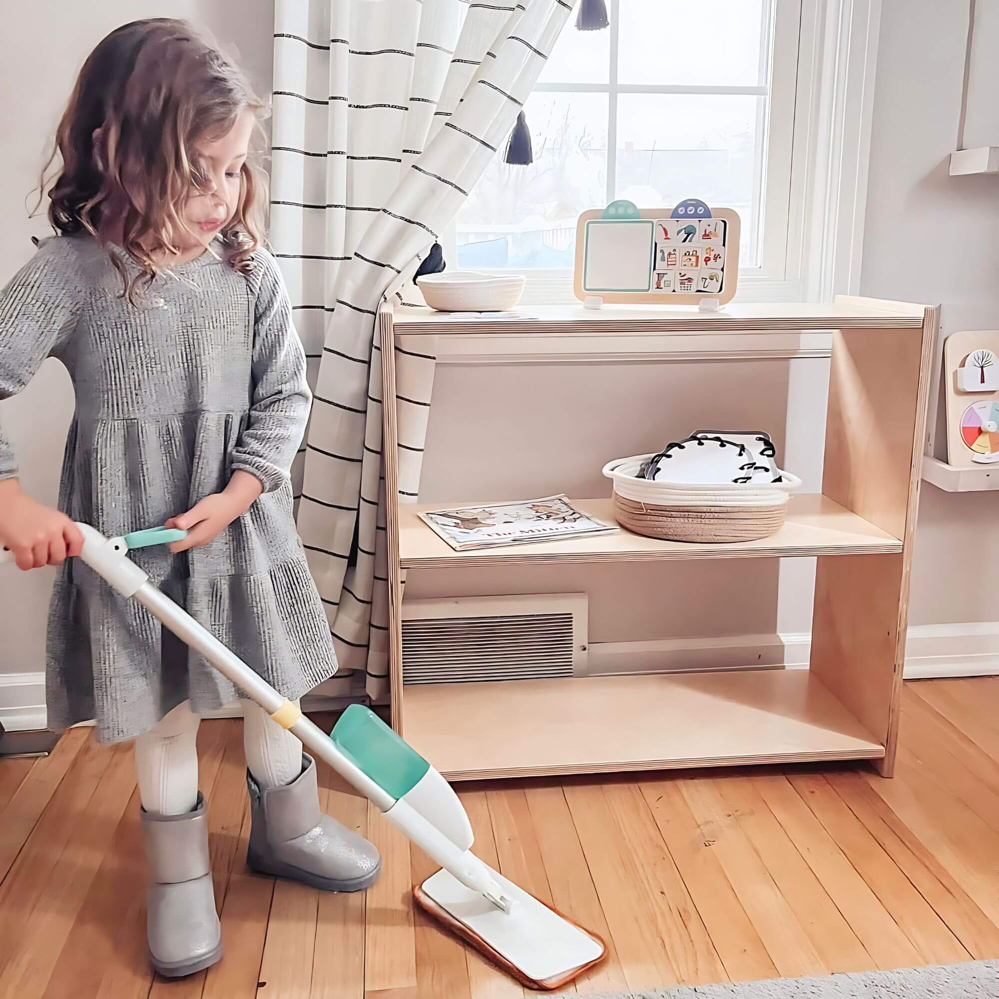 Child using a mop in a room with a wooden floor and a shelf.