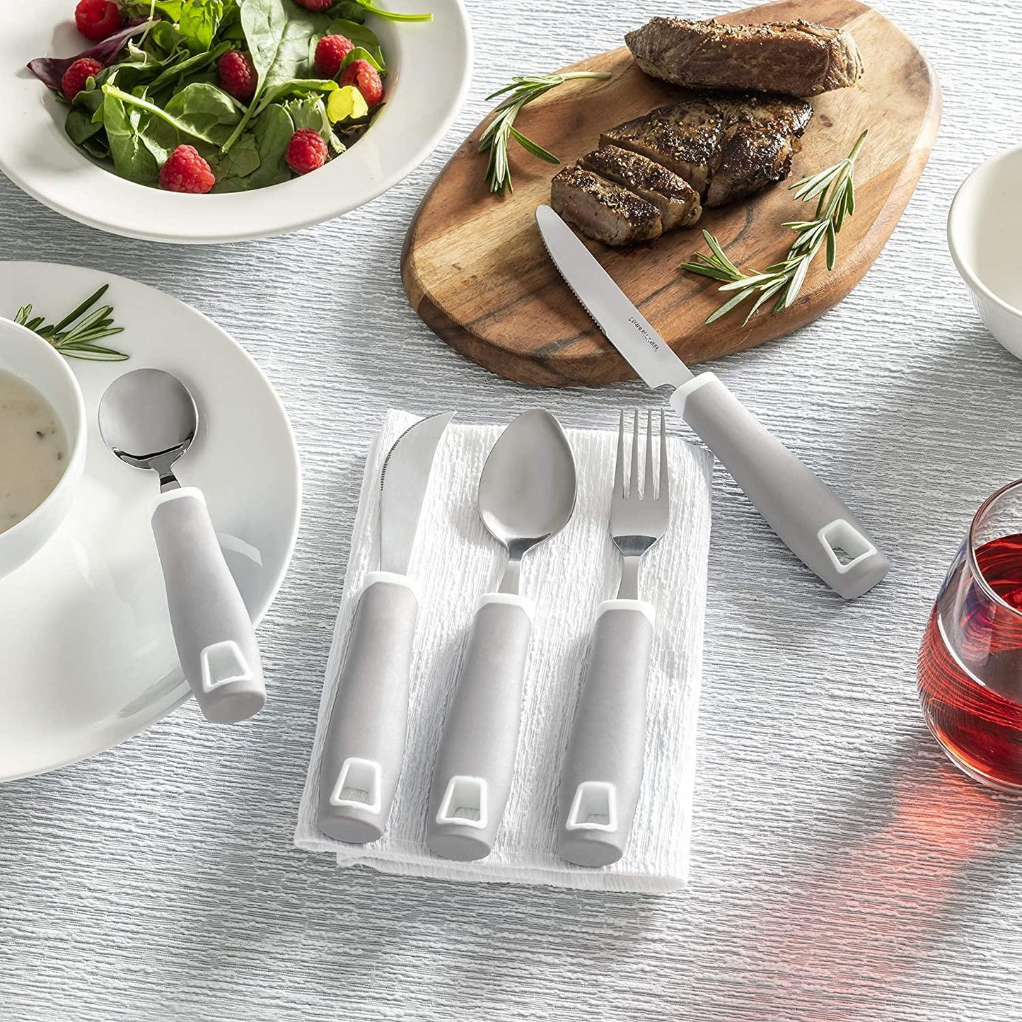 Adaptive utensils with thick gray handles on a table set with salad, steak, soup, and a drink
