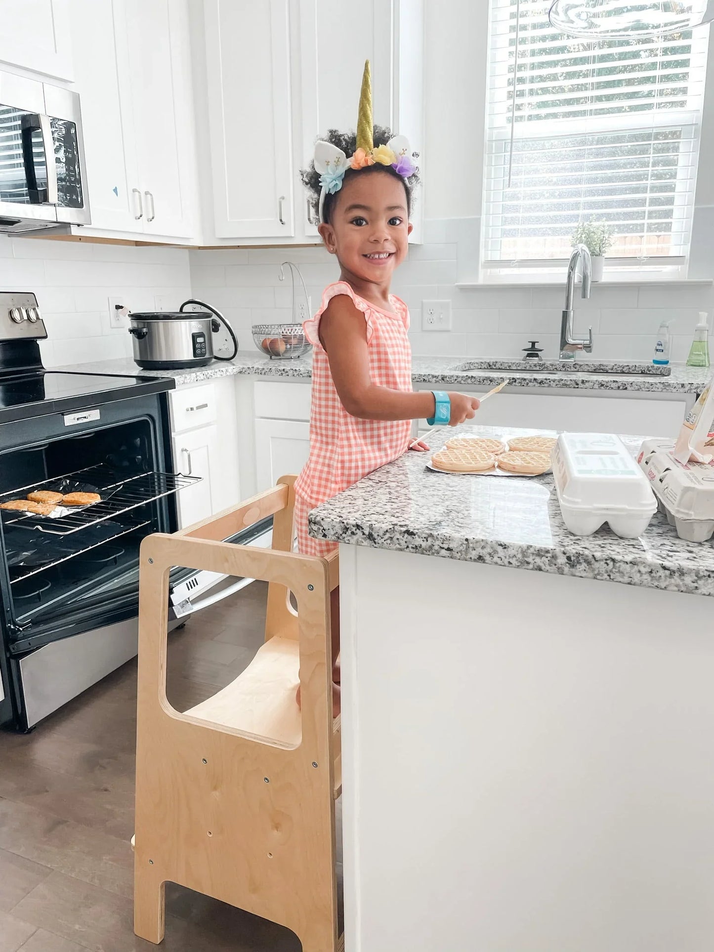 Child baking in kitchen on wooden learning tower, wearing unicorn headband, sensory-friendly