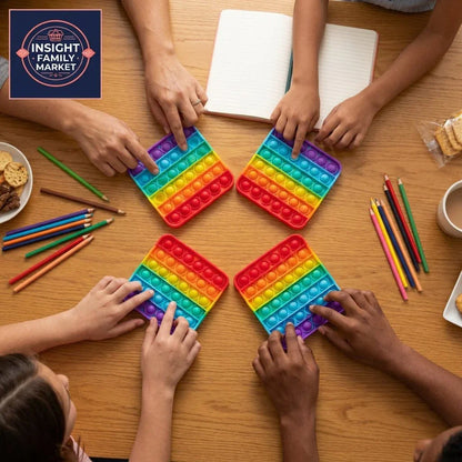 Family hands playing with rainbow pop fidget toys at a wooden table with colored pencils.
