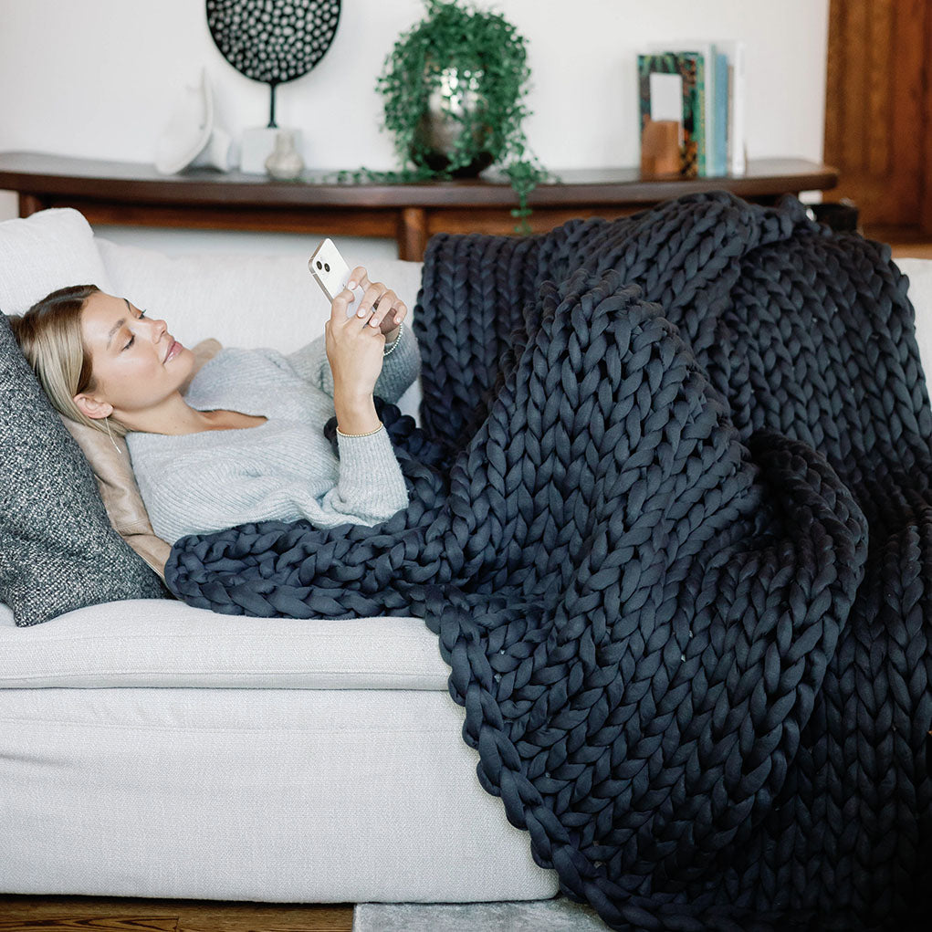 Woman lying on a couch with a dark gray knit blanket, using a smartphone.