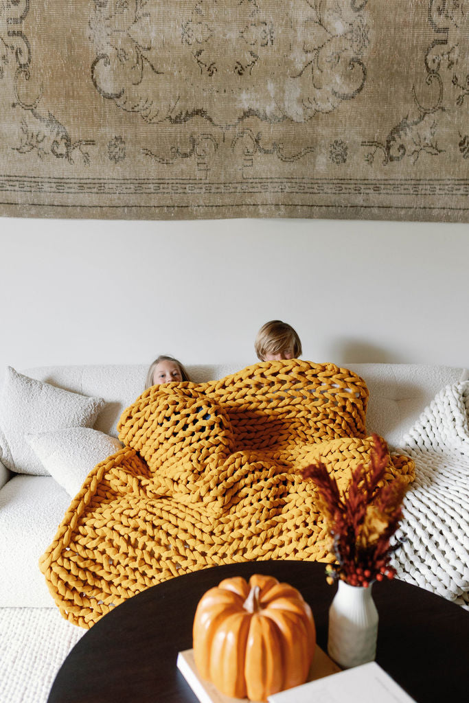Two children under a mustard yellow blanket on a couch with a pumpkin and vase on a table in front.