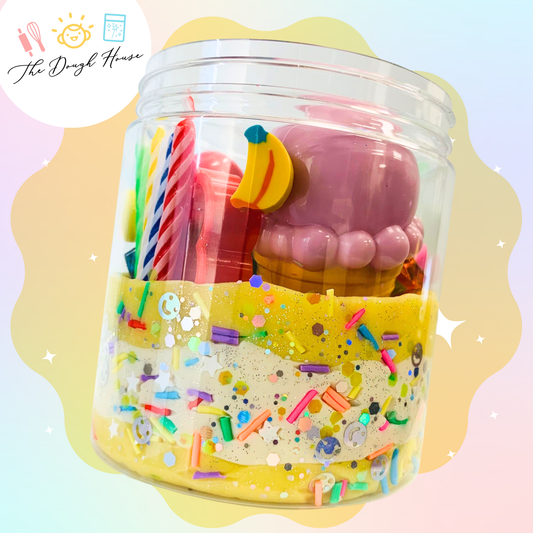 Clear jar with colorful contents including a pink ice cream, banana, and candles on a light purple background.