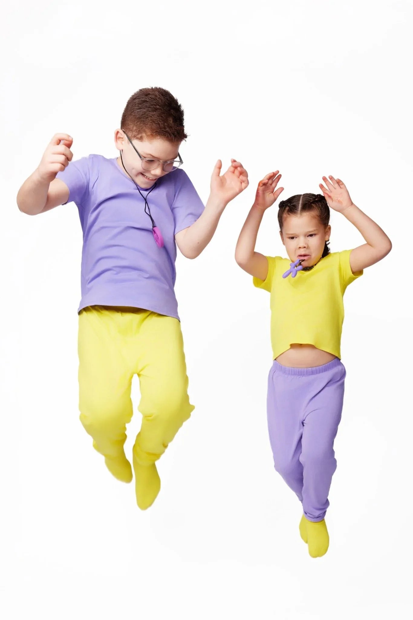 Two kids jumping in sensory-friendly clothing, one wearing chewelry, on a white background.