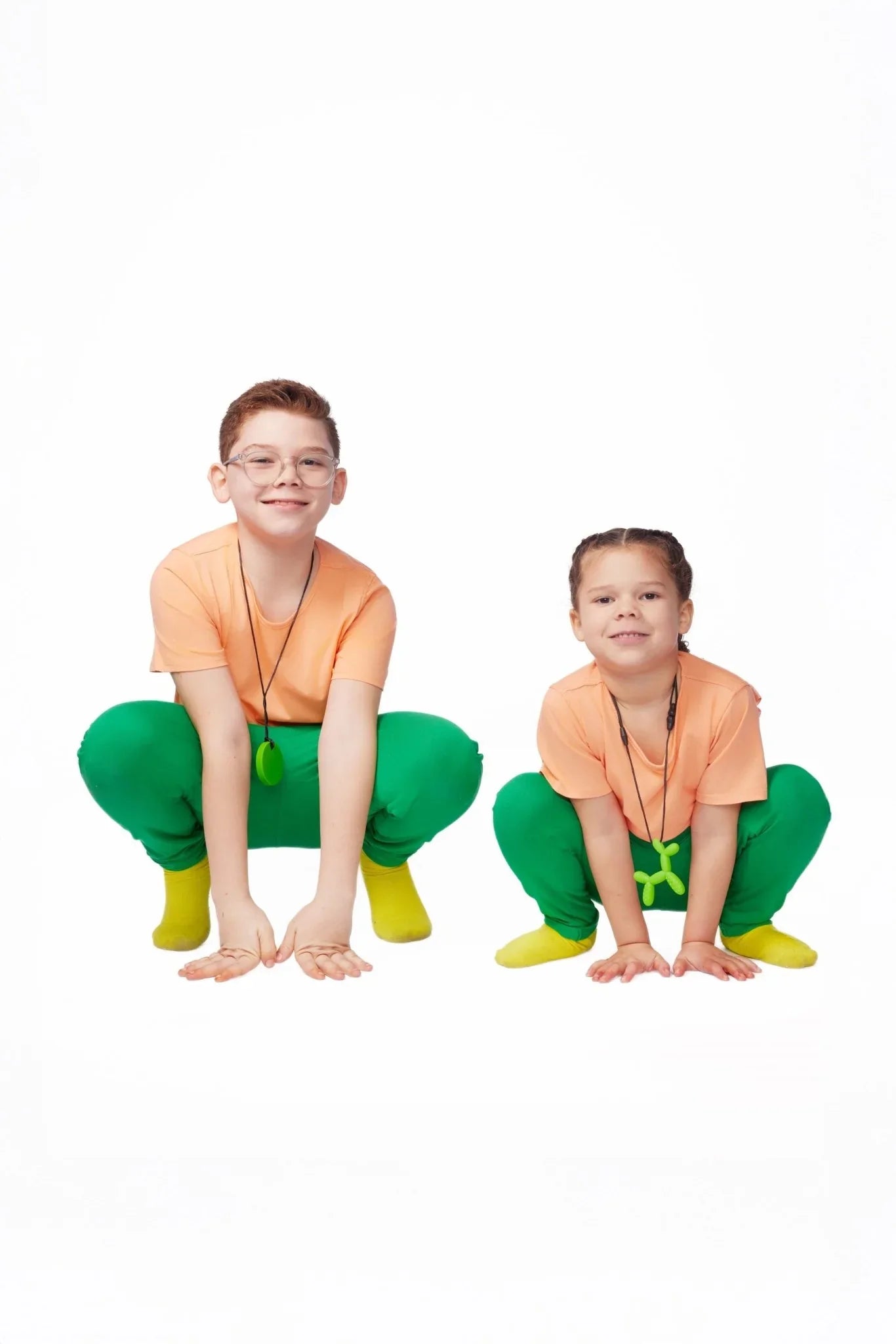 Two kids in sensory-friendly orange shirts, green pants, and yellow socks squatting and smiling