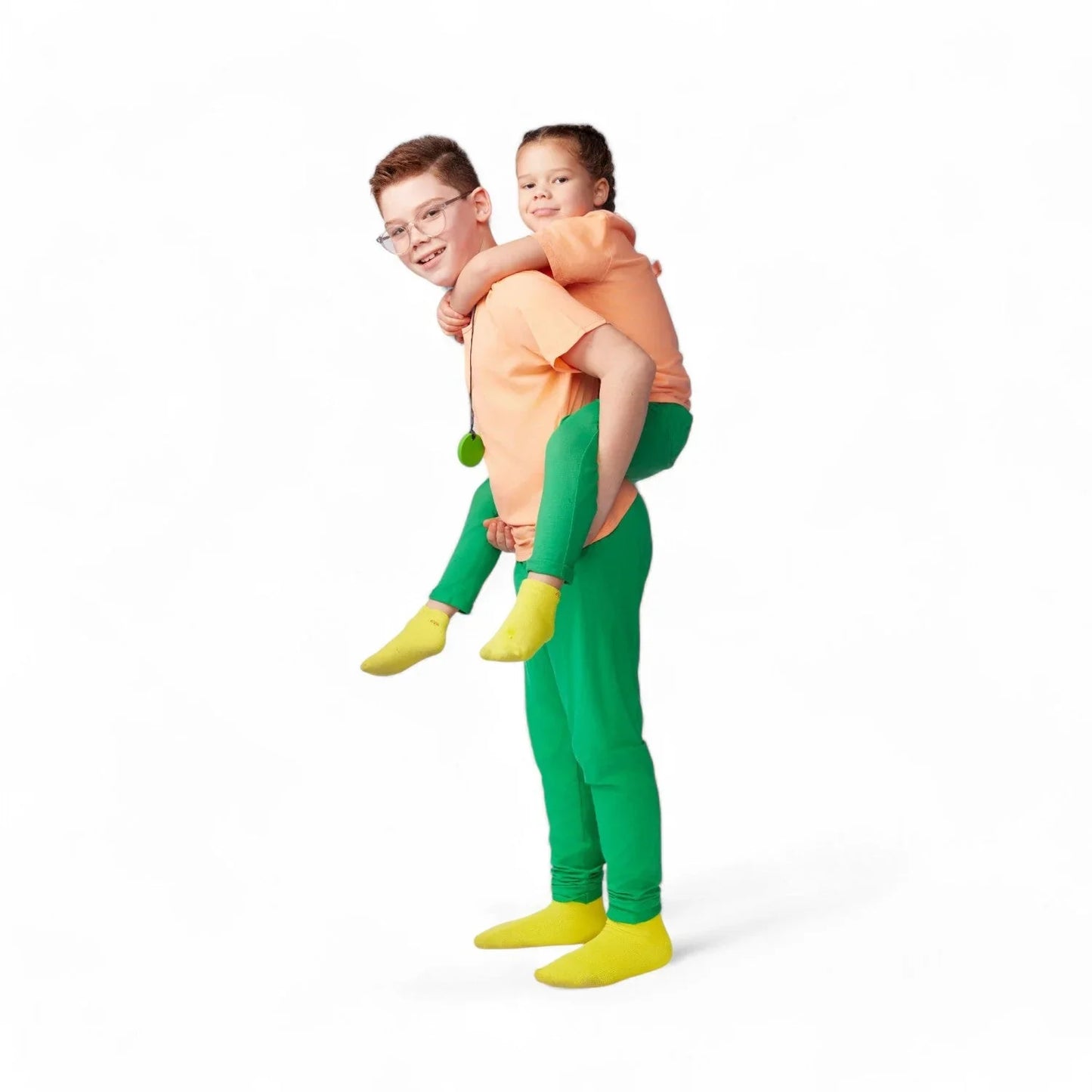 Two children in sensory-friendly clothing, piggyback pose, orange shirts, green pants, yellow socks