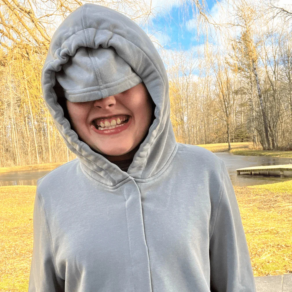 Smiling child in sensory-friendly gray hoodie with hood up outdoors near trees and pond