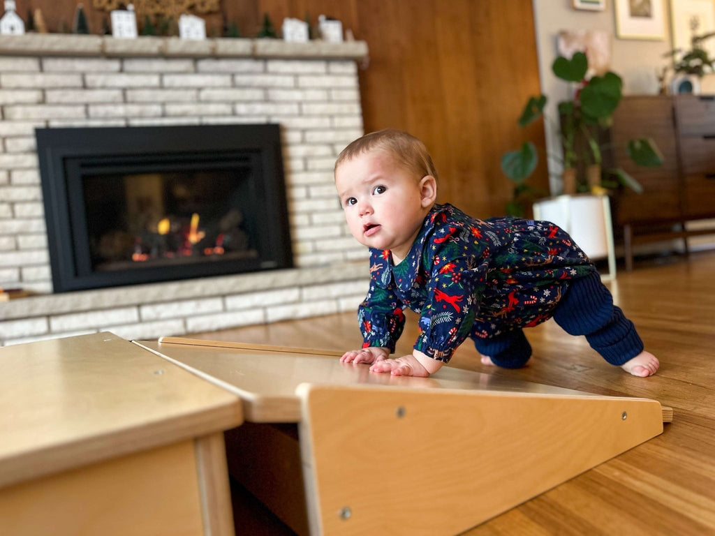 Baby in a colorful onesie crawling on a wooden play structure in a living room with a fireplace.