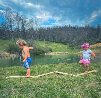 Two young children balancing barefoot on a zigzag wooden balance beam outdoors by a pond