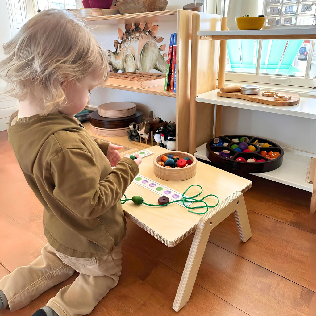 Child playing with educational toys at a wooden table in a room with shelves and windows.