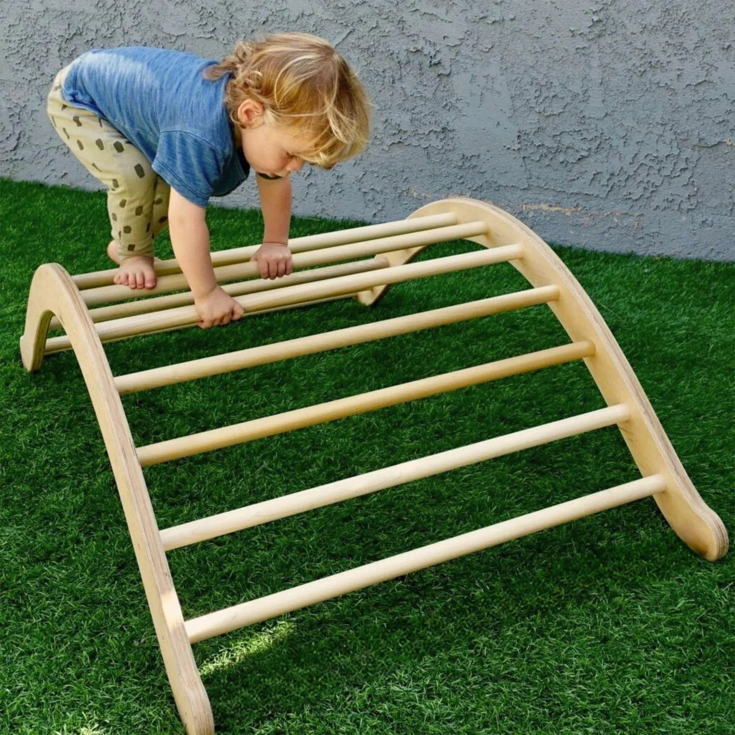 Toddler climbing on a wooden Pikler arch on artificial grass outdoors
