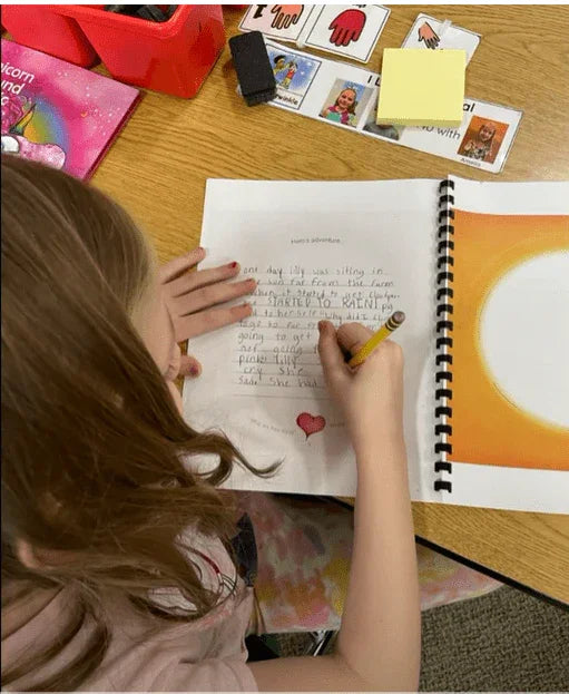 Child writing in a spiral-bound blank book with sensory-friendly tools on table