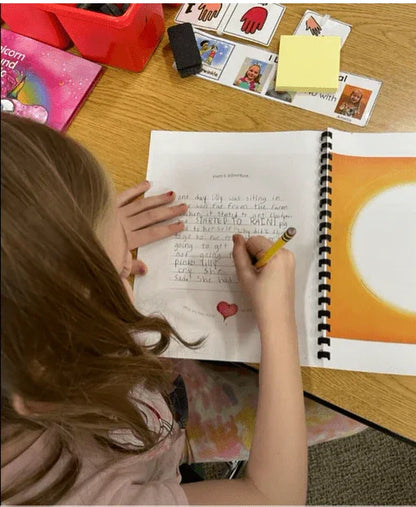 Child writing in a spiral-bound blank book with sensory-friendly tools on table