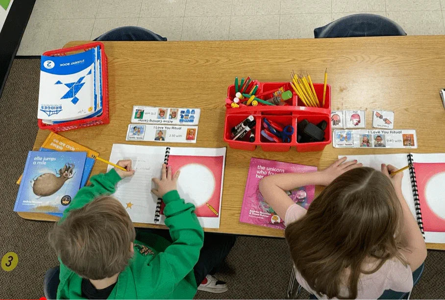 Two children drawing in spiral workbooks at classroom table with learning supplies and books