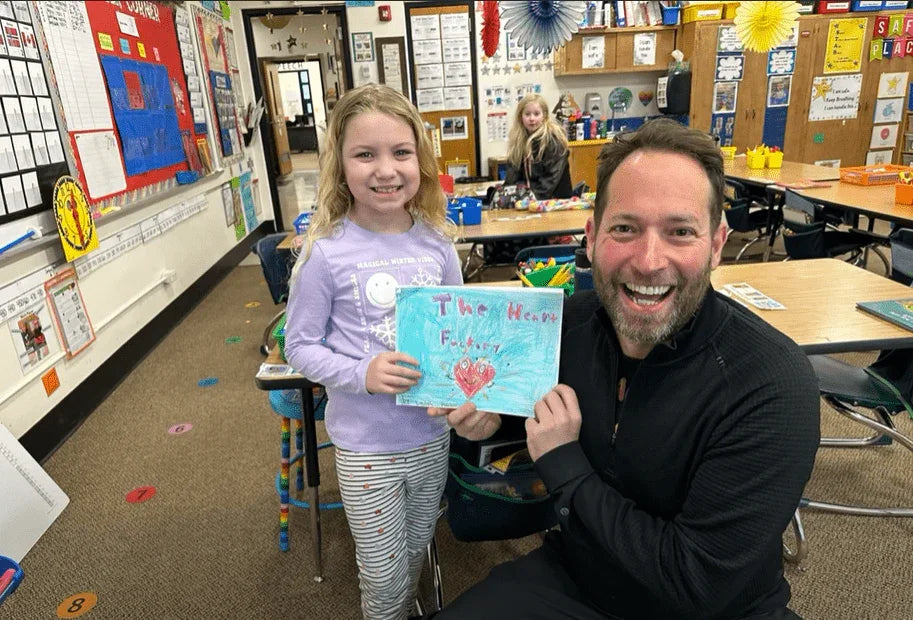 Girl and man smiling in a classroom holding a handmade book titled 'The Heart Factory'.