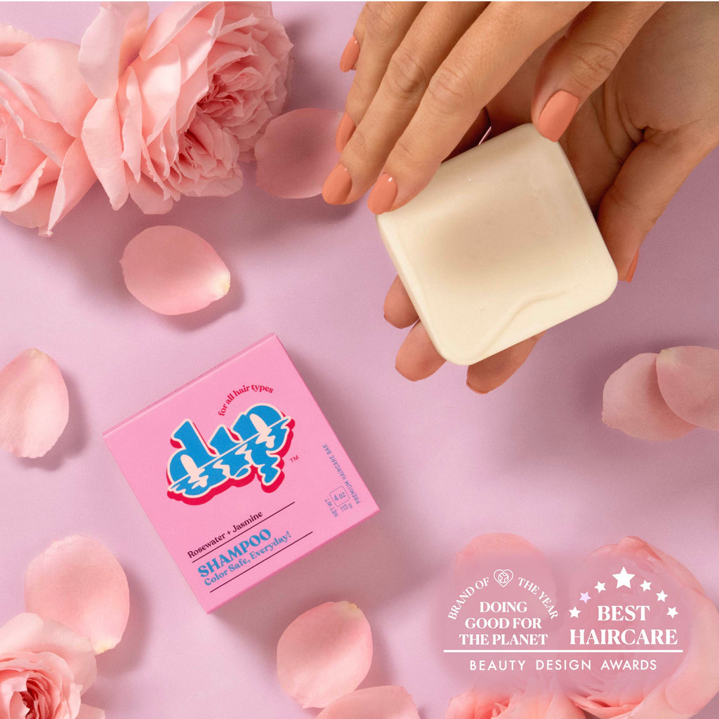 Hand holding a bar of soap with a pink shampoo box and roses on a pink background