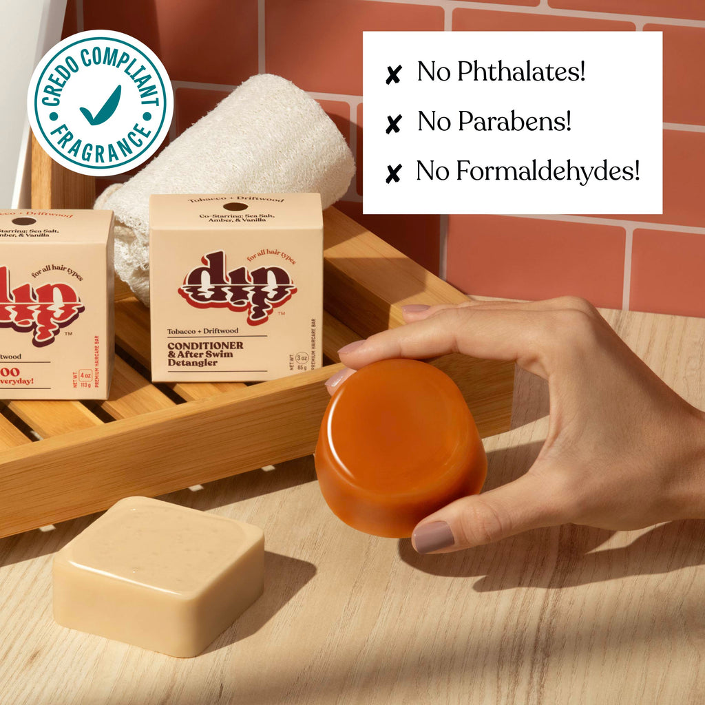 Hand holding a jar of soap next to soap bars and boxes on a wooden shelf with a brick wall background.
