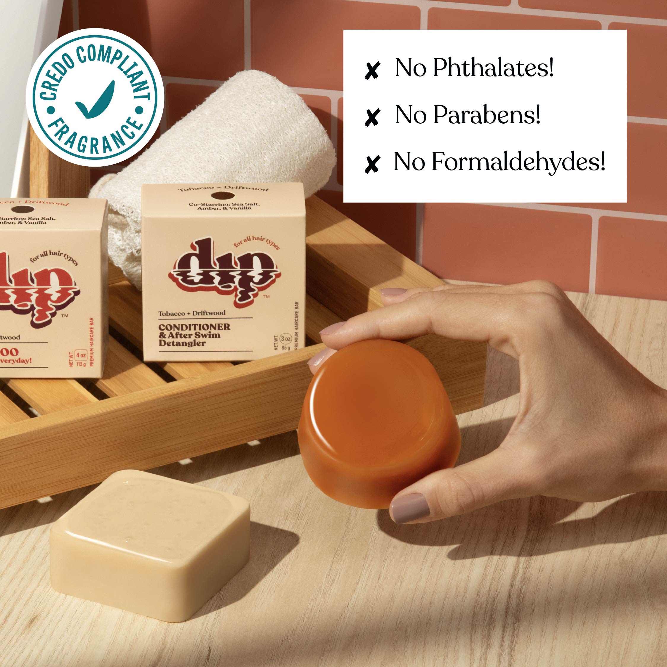 Hand holding a jar of soap next to soap bars and boxes on a wooden shelf with a brick wall background.