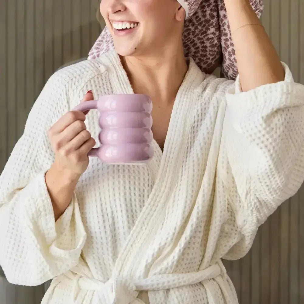 Woman in a white waffle robe holding a pink mug, smiling, with hair wrapped in a towel.
