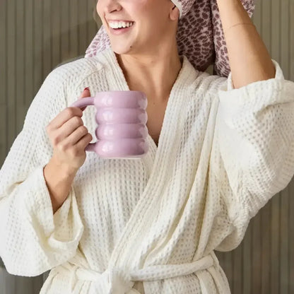 Woman in a white waffle robe holding a pink mug, smiling, with hair wrapped in a towel.