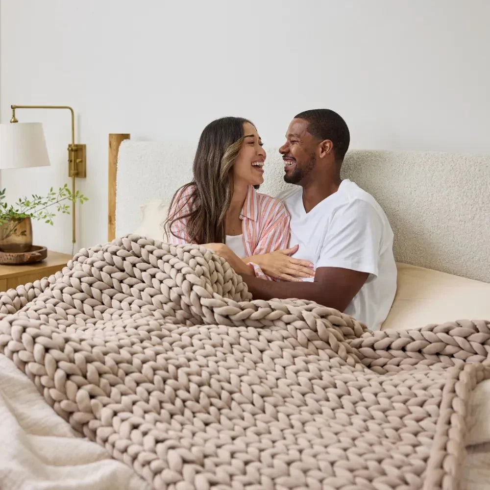 Couple sitting on bed under chunky knit cooling weighted blanket in neutral coastal sand color