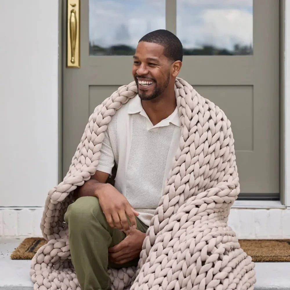 Man sitting outside wrapped in a beige chunky knit weighted blanket, smiling.