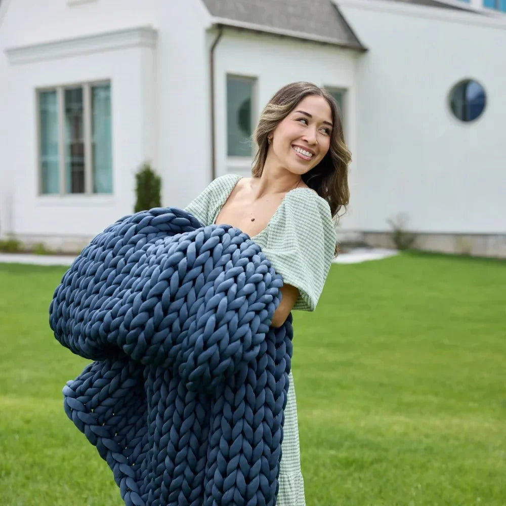 Smiling woman holding blue chunky knit weighted blanket outdoors on green lawn