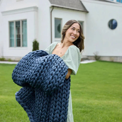 Smiling woman holding blue chunky knit weighted blanket outdoors on green lawn
