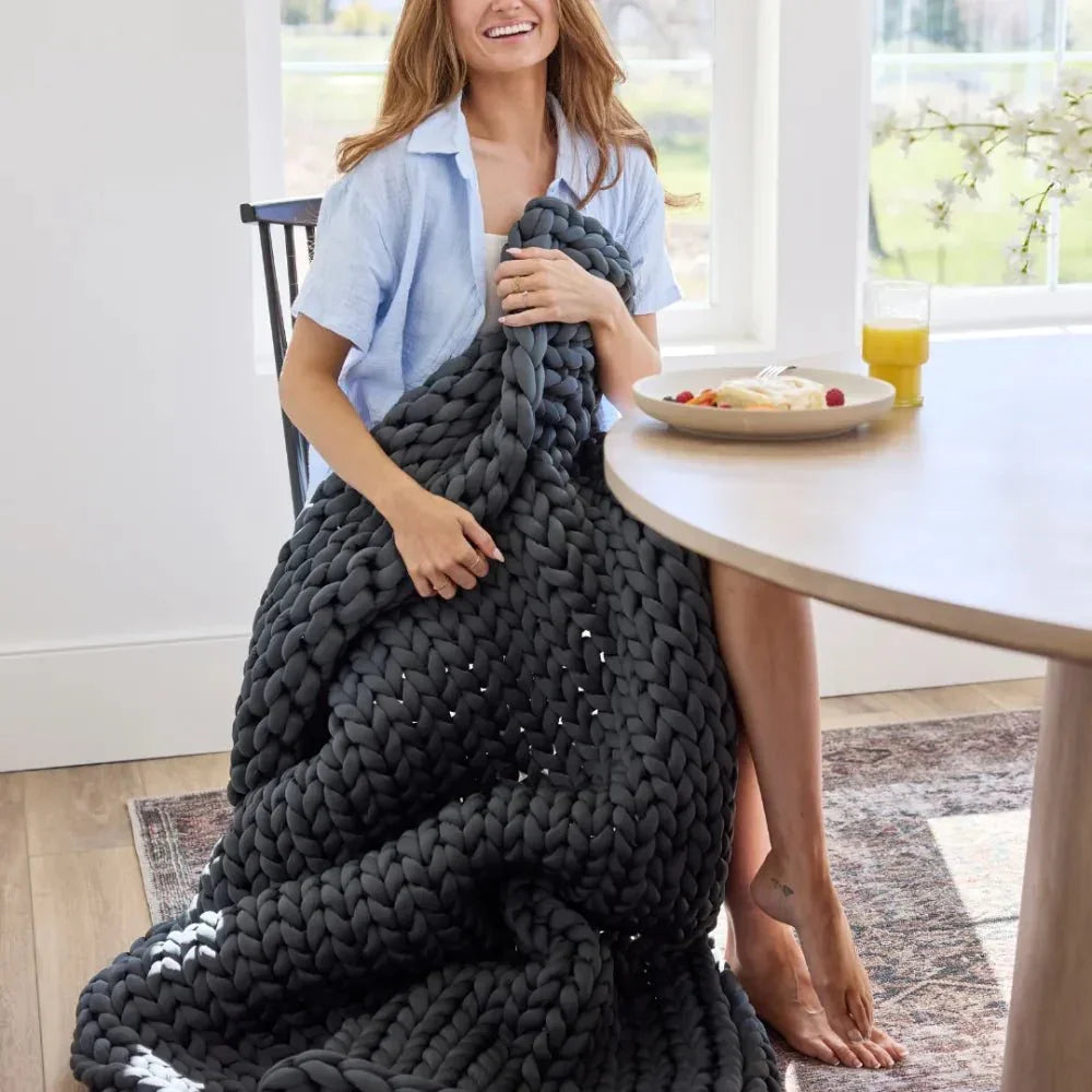 Woman sitting at table with chunky knit grey weighted blanket and breakfast, bright room