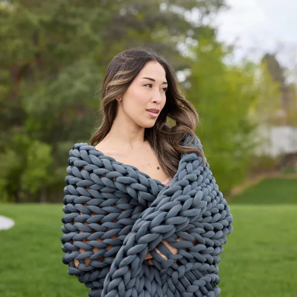 Woman wrapped in chunky knit grey weighted blanket outdoors on a green lawn