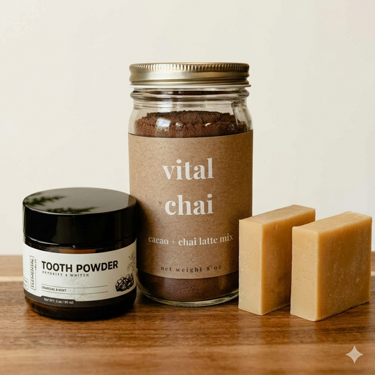 A jar of 'vital chai' cacao + chai latte mix, a small jar of tooth powder, and two bars of soap arranged on a wooden surface against a light beige background.