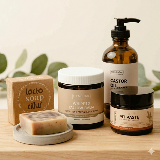 A set of natural skincare products from Elemental on a light wood surface with a soft, neutral background and blurred green leaves. The products include a boxed Lactoo Soap Cillus, a bar of soap on a small dish, a Whipped Tallow Balm jar, a Castor Oil bottle, and a Pit Paste jar.